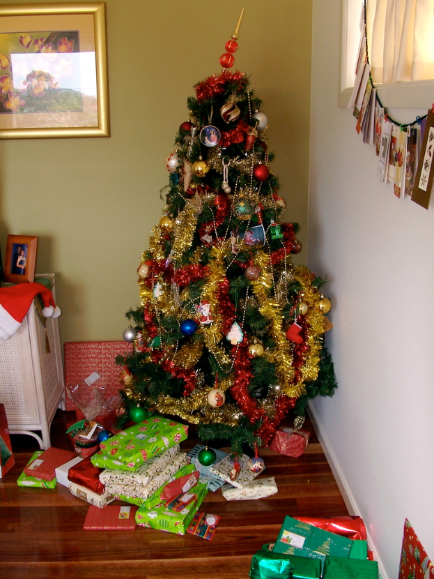 Ein geschmückter Weihnachtsbaum mit Geschenken am Boden, ein Foto Rahmen an der linken Wand und ein Vorhang am rechten Fenster.