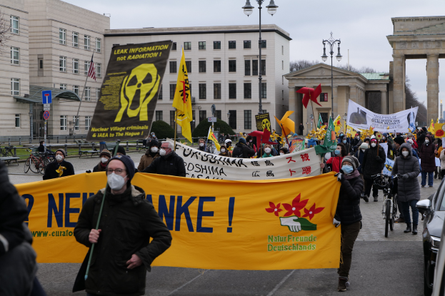 Eine große Gruppe von Menschen marschiert mit Schildern und Fahnen auf einer Straße bei einer Anti-Atom-Demonstration in Deutschland, mit Fahrzeugen auf der rechten Seite und Gebäuden, Laternen, Bäumen und einem Tor im Hintergrund.