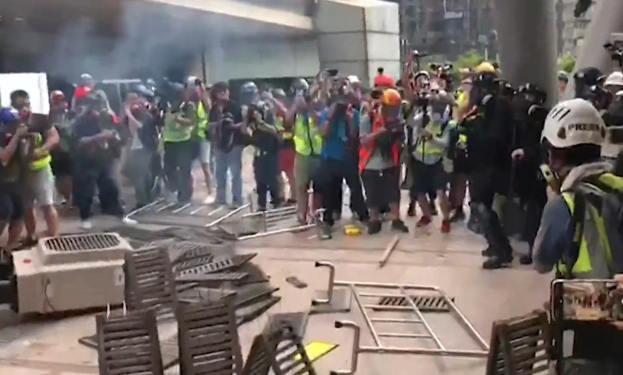 Menschen in Helmen und Jacken stehen vor einem rauchenden Gebäude, einige halten Kameras, mit verstreuten Metallobjekten und Stadtgebäuden im Hintergrund.