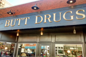 Außensicht eines Gebäudes mit einer Leuchtschrift "butt drugs", das von Glaswänden und -türen umgeben ist, mit Fahrzeugen, Gebäuden, Bäumen und Himmel im Hintergrund.