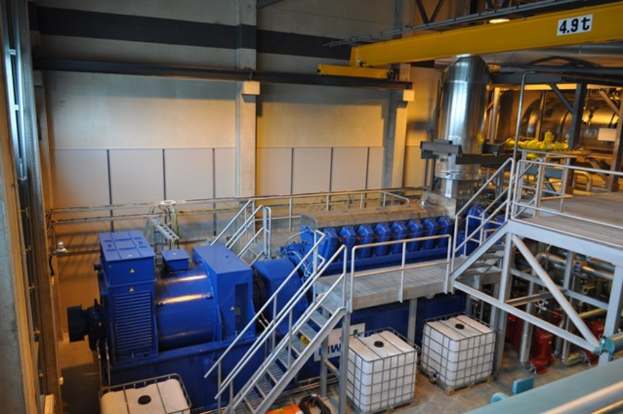 Eine Fabrikinnenszene mit Trägern, einer Treppe, Geländer, Containern, Lampen, Rohren und Wänden.