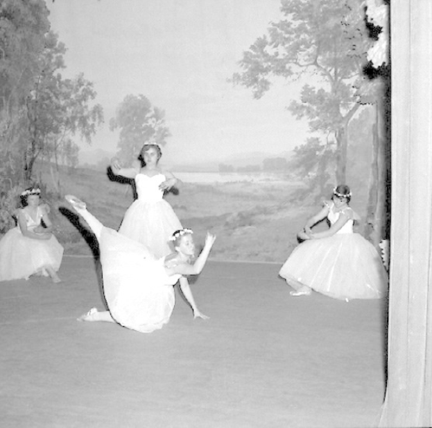 Vier junge Ballerinen in weißen Kleidern tanzen auf einer Bühne mit einer gemalten Kulisse aus Bäumen und Himmel in Schwarz-Weiß.