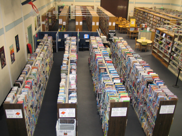 Ein Bibliotheksinneres mit Bücherregalen, die mit Büchern gefüllt sind, und Tischen und Stühlen, die vor einer Wand angeordnet sind.