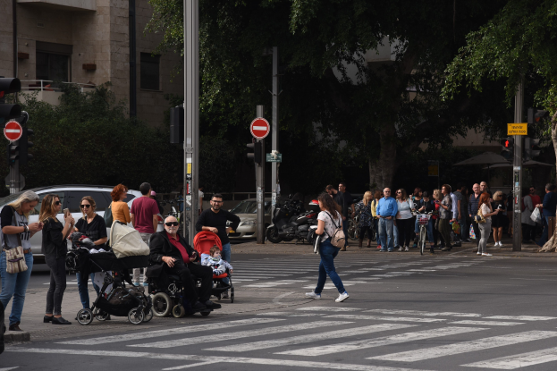 Gruppe von Menschen, die eine Straße an einer Ampel überqueren, einige mit Taschen, mit einer Person in einem Kinderwagen; Fahrzeuge, Verkehrszeichen, Schilder, Bäume und Gebäude im Hintergrund.