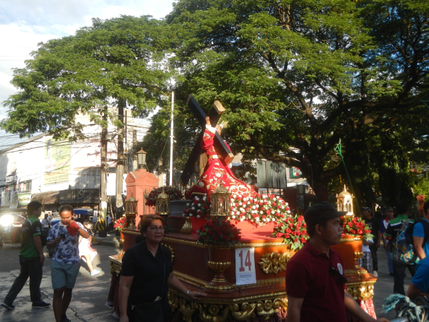 Eine Gruppe von Menschen, die in einer Prozession an einer Jesus-Statue vorbeigehen, mit Bäumen, Gebäuden, Strommasten und Fahrzeugen im Hintergrund unter einem bewölkten Himmel.