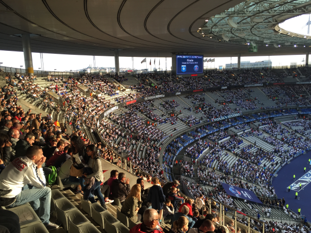 Eine große Menschenmenge sitzt im Allianz Arena Stadion in München, Deutschland, und schaut ein Fußballspiel, mit einer Bühne auf der rechten Seite, Fahnen, Stangen und einem Bildschirm im Hintergrund, und der Himmel ist oben sichtbar.