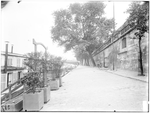 Ein Schwarz-Weiß-Foto einer Straße mit einer Wand auf der rechten Seite, Bäumen und Pflanzen auf der linken Seite und einer Brücke im Hintergrund, mit Text unten, der lautet: "New York City, New York, New Jersey, NY, USA, USA - Straßenansicht des New Yorker Staatsgefängnisses".