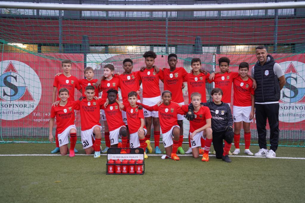 Eine Gruppe junger Männer in roten T-Shirts und schwarzen Shorts steht auf einem Fußballfeld vor einer Kiste roter Gegenstände, im Hintergrund Schilder mit der Aufschrift "FC Red Black Eglaute - Champions League".