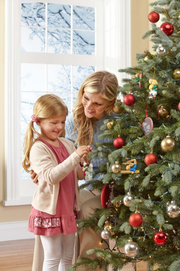 Ein geschmückter Weihnachtsbaum mit einer Frau und einem Kind davor, ein Fenster und eine Wand im Hintergrund und sichtbare Tannenbäume außerhalb des Fensters.