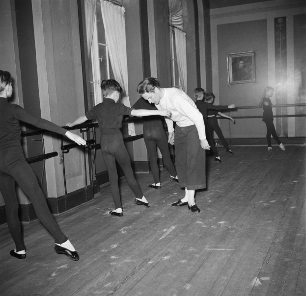 Eine Gruppe junger Tänzer in einem Tanzstudio, die vor einer Wand mit einem gerahmten Schwarz-Weiß-Foto eines Choreografen stehen, mit Fenstern und Vorhängen im Hintergrund.