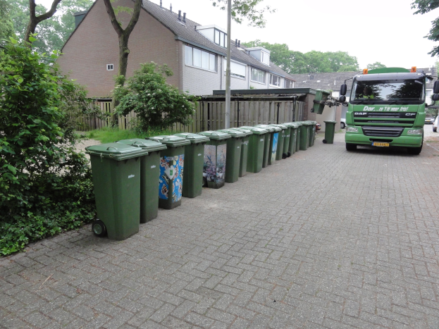 Ein grüner Lastwagen neben einer Reihe grüner Mülltonnen auf einer Straße, mit Pflanzen und Bäumen links und Gebäuden, einem Mast, einem Zaun und Himmel im Hintergrund.