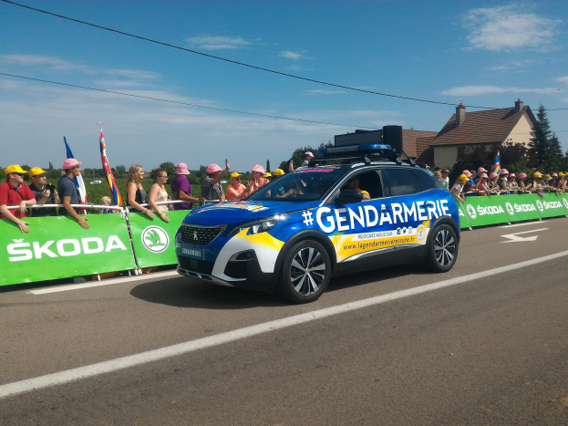 Ein Polizeiwagen fährt eine Straße entlang neben einer Menschenmenge, einige tragen Mützen und halten Fahnen, mit einem Banner auf dem Wagen, Bäumen, Gebäuden und einem bewölkten Himmel im Hintergrund, und Text am unteren Rand, der 'Gendarmerie de la Tour de France' lautet.