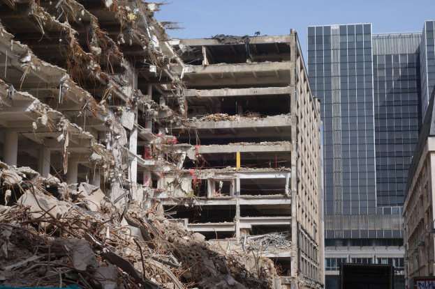Ein großes Gebäude wird inmitten einer Stadt abgerissen, umgeben von intakten Gebäuden, Straßeninfrastruktur, Fahrzeugen, Bäumen und einem klaren blauen Himmel.