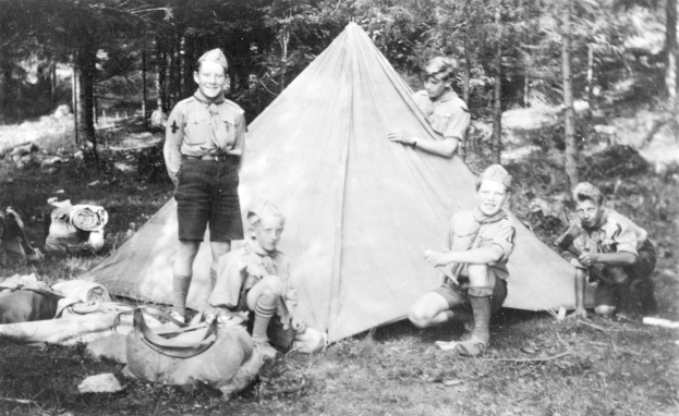 Schwarzes und weißes Bild von Pfadfindern in Uniformen, die sich um ein Zelt in einem bewaldeten Gebiet versammelt haben, einige sitzen und andere stehen, mit verstreuten Taschen und Gegenständen.