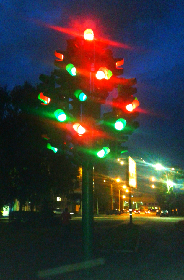 Eine nächtliche Ampel mit grünen und roten Lichtern, umgeben von Fahrzeugen, Bäumen, Gebäuden und Lichtern im Hintergrund unter dem Nachthimmel.