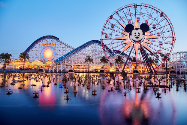 Eine lebendige Disneyland California Adventure Szenerie bei Sonnenuntergang mit einem riesigen Mickey-Maus-Rad im Vordergrund, einer Achterbahn im Hintergrund und Booten auf dem Wasser, mit einem Himmel aus Orangen, Pinks und Purpurs und silhouettenartigen Bäumen.