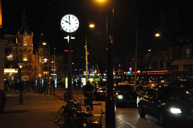 Eine nächtliche Straßenansicht mit Fahrzeugen auf der Straße, Fahrrädern neben Pfählen, einer Uhr an einem Pfahl, beleuchteten Gebäuden im Hintergrund und Menschen auf dem linken Gehweg.