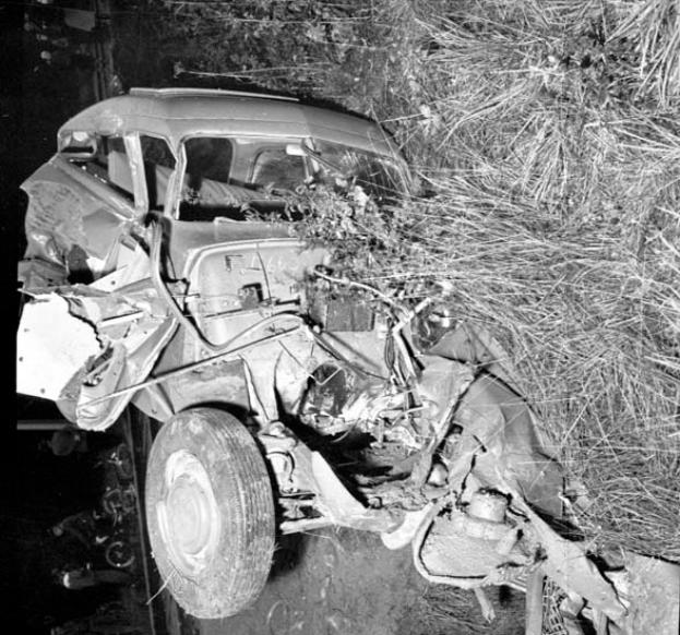 Ein Schwarz-Weiß- Foto eines schwer beschädigten Autos am Straßenrand mit einer zerbeulten Front, zersplitterten Fenstern, einem Baum im Hintergrund und verstreuten Gegenständen um die Unfallstelle.