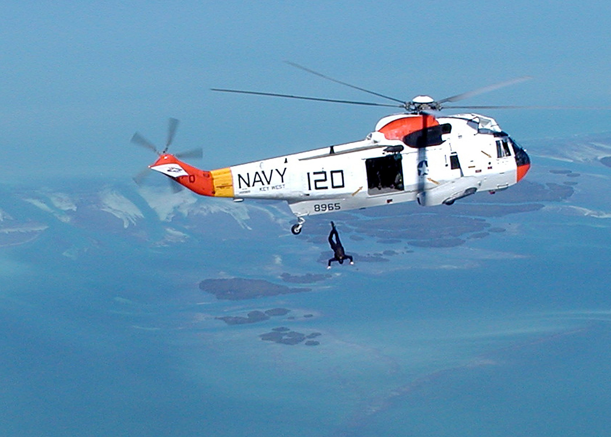 Ein Rettungshubschrauber fliegt über Wasser mit einer Person darunter, vor einem Himmel-Hintergrund.