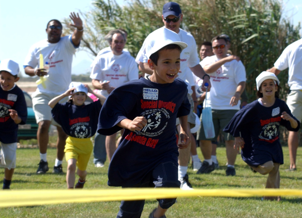 Kinder laufen in einem Wettbewerb auf einem grünen Rasen mit einem gelben Band im Vordergrund, einige tragen Kappen und Schutzbrillen, andere halten Gegenstände, vor einem Hintergrund aus Bäumen und einem klaren blauen Himmel.