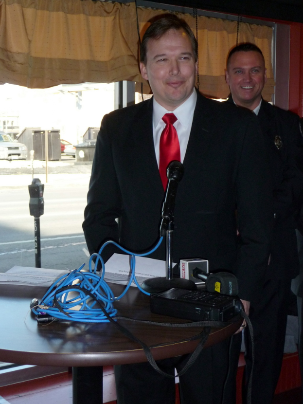 Eine Person in einem Anzug lächelt und steht vor einem Tisch mit Mikrofonen, Kabeln und Dokumenten, während eine andere Person hinter ihr steht, mit einem Blick auf Fahrzeuge, Pfosten und eine Straße durch ein Glasfenster mit einem Vorhang.