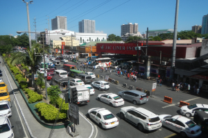 Eine belebte Stadtstraße mit Autos, Bussen, Fußgängern, Bäumen und Gebäuden unter einem klaren blauen Himmel.