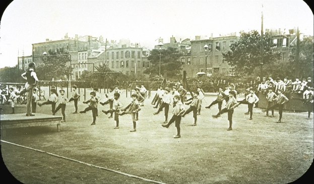Schwarzes Foto von Kindern, die auf einem grasigen Fuballfeld mit Baumen, Masten und Gebauden mit Fenstern im Hintergrund Fuball spielen.