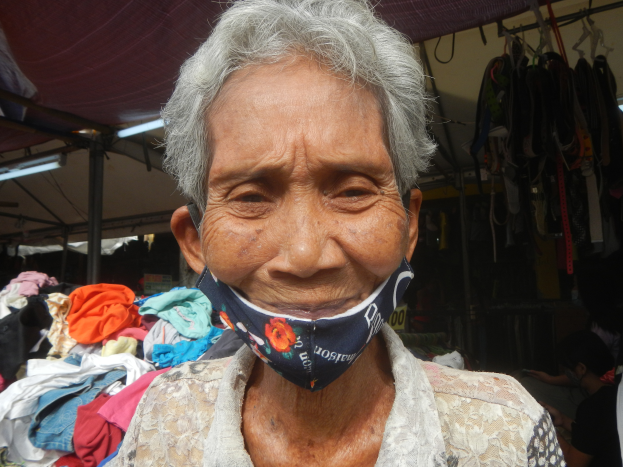Eine ältere Frau mit Gesichtsmaske steht auf einem Markt umgeben von Körben und Lichtern, mit einer weiteren Person im Hintergrund.