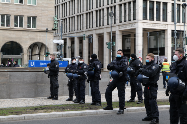 Eine Gruppe von Polizisten in schwarzen Uniformen und Masken steht vor einem Gebäude mit Glasfenstern und Säulen in Berlin, Deutschland, einige halten Helme, mit Laternen, Verkehrsampeln, Schildern mit Text, einer Statue und anderen Menschen im Hintergrund auf einem grasbewachsenen Boden.