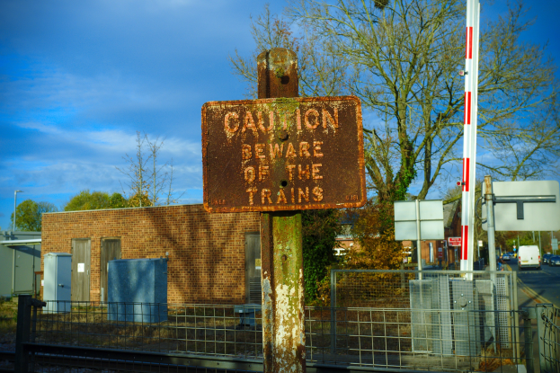 Vorsichtsschild an einem Bahnschienenzaun neben Bäumen, Pfählen, einem Gebäude mit Fenstern, Containern, einem Straßenmast, Fahrzeugen auf der Straße und einem bewölkten Himmel.