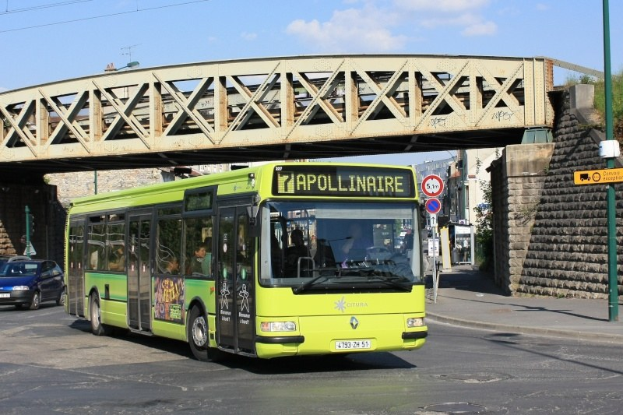Grüner Bus fährt unter einer Brücke auf einer Stadtstraße mit anderen Fahrzeugen, einem Mast mit Schildern, einer Wand, Gebäuden, Bäumen und einem bewölkten Himmel.