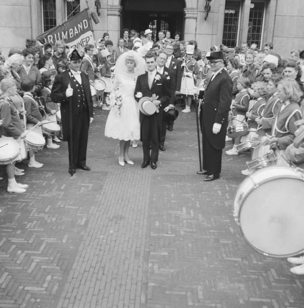 Eine Braut in einem weißen Kleid und Schleier und ein Bräutigam mit einem Blumenstrauß gehen eine Straße entlang vor einer Menge, einige mit Musikinstrumenten, mit einem Gebäude und einem Banner im Hintergrund, alles in Schwarz-Weiß.