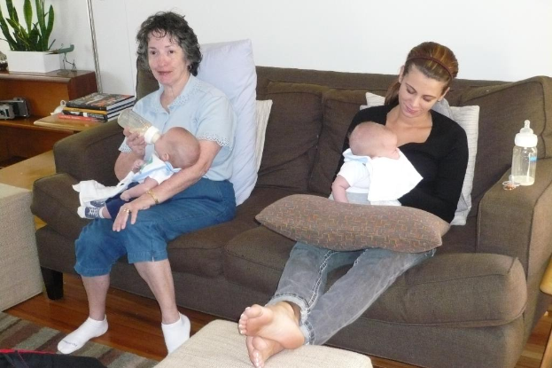 Zwei Frauen sitzen auf einer Couch und halten Babys, mit Kissen dazwischen, vor einer weißen Wand, mit einem Schreibtisch, Büchern und einer Zimmerpflanze links.