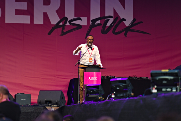 Ein Mann in einem weißen Hemd und braunen Hosen steht auf einer Bühne und hält ein Mikrofon, mit einem pinken Banner im Hintergrund, auf dem 'Berlin as Fuck' steht, und Lautsprechern und anderen Gegenständen auf der Bühne.