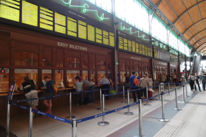 Menschen stehen in einer Schlange an einem Bahnhof mit Taschen, Stangen mit Bändern, Informationsschildern, Deckenleuchten und Treppen mit Gelöndern, mit einem Gebäude im Hintergrund.