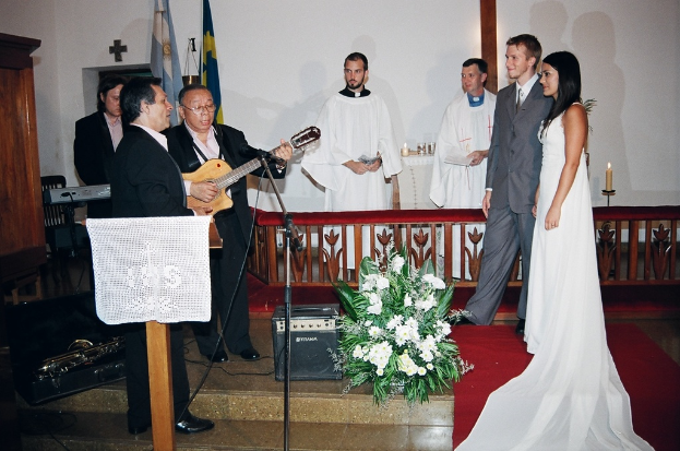 Eine Hochzeitszene in einem Raum mit zwei Pastoren, einer Braut, einem Bräutigam und drei Musikern; ein Podium mit Stoff vor den Musikern; ein Tisch mit Kerzen hinter den Pastoren; ein Kerzenhalter hinter dem Paar; ein Bukett, Lautsprecher, Taschen und ein roter Teppich auf dem Boden; sowie Flaggen, eine Wand mit einem Kreuz, einen Schrank und einen Stuhl im Hintergrund.