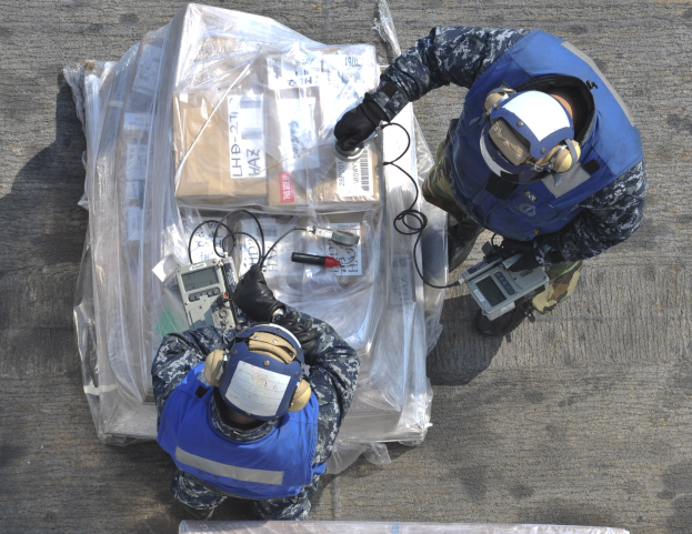 Zwei Männer in blauen Uniformen und Helmen arbeiten an einem großen Stück Ausrüstung, das wie eine Drohne aussieht, während sie verschiedene Gegenstände halten, mit einer Plastikabdeckung auf dem Boden.