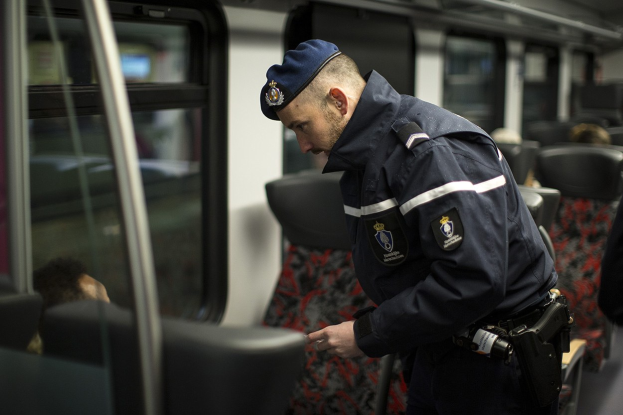 Polizist in Uniform und Mütze auf einem Zug stehend, der auf sein Handy schaut, mit sitzenden Passagieren und einer weiteren Person im Bild.