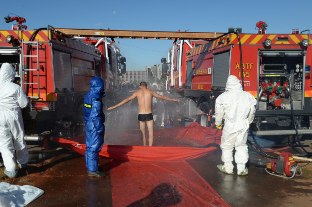 Drei Personen in Warnwesten stehen im Vordergrund, wobei eine Person auf einer Abdeckung mit Wasser davor steht. Einige andere sind in der Nähe, und Feuerwehrautos mit ausgefahrenen Metallstangenleitern sind daneben positioniert. Gebäude sind im Hintergrund sichtbar, und es gibt eine Abdeckung mit einigen Gegenständen auf der Oberfläche.