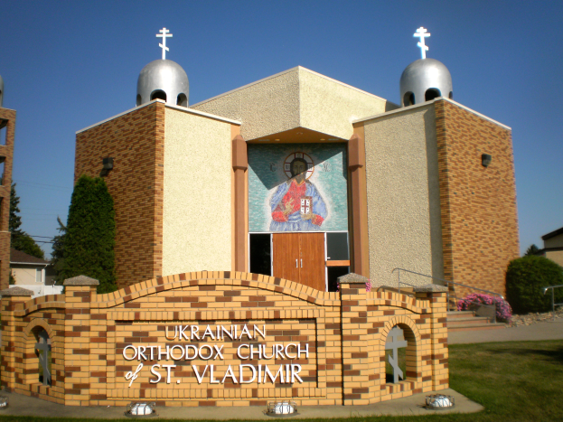 Die Ukrainische Orthodoxe Kirche St. Wladimir mit einer texttragenden Wand, Gras, Gebäuden, Bäumen, blühenden Pflanzen, Treppen mit Geländern und einem Wandgemälde unter einem sichtbaren Himmel.