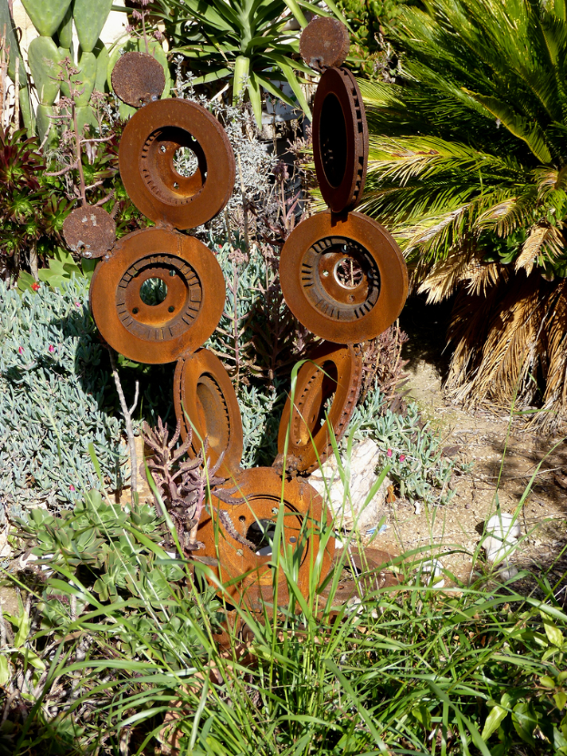 Eine Metallskulptur steht auf einer Landschaft mit spärlicher Vegetation, einschließlich einiger Pflanzen, Grasflecken und verstreuten Bäumen.