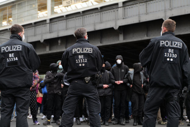 Polizeibeamte in schwarzen Uniformen und Masken stehen vor einer Menge bei einer Demonstration in einer städtischen Umgebung mit einer Brücke und einem Gebäude im Hintergrund.