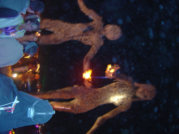 Eine Gruppe von Menschen vor brennenden Statuen mit Lichtern im Hintergrund.