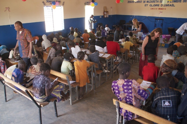 Kinder auf Bänken in der Nähe von Erwachsenen sitzend, während Stühle, eine Tür, ein Fenster, ein Tisch, Luftballons und Text im Hintergrund zu sehen sind.