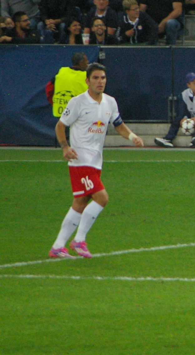 Ein Mann in einem weißen Hemd und roten Shorts, der auf einem Feld Füßball spielt, mit einer Gruppe von Menschen im Hintergrund.