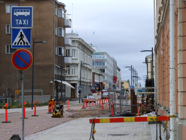 Stadtstraße mit Gebäuden, Straßenlaternen, Schildern, Verkehrszeichen, Baustellenabsperrungen, Fahrzeugen, Absperrpoller, Bäumen und einer Baustelle mit Verkehrszeichen unter einem bewölkten Himmel.