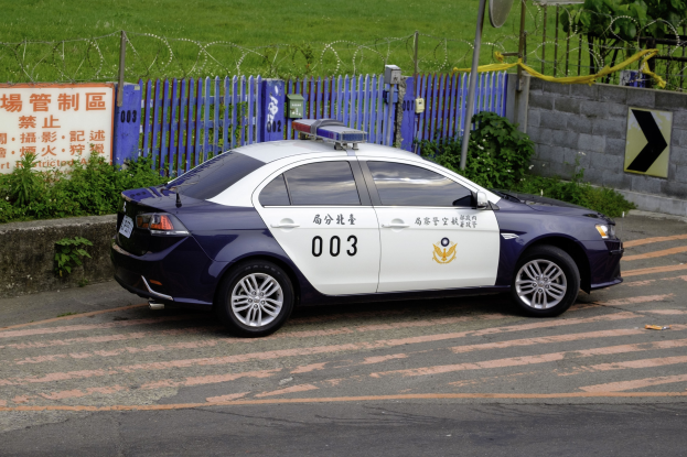 Polizeiauto am Straßenrand neben einem Zaun, einer Tafel mit Text, einer Wand, einem Pfahl, einer Schüsselantenne und Bäumen.