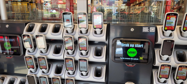 Ein Verkaufsautomat mit mehreren Handys auf der Oberseite, vor einem belebten Hintergrund aus Menschen, Geschäften, Schildern und Lichtern.