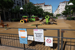 Ein umzäunter städtischer Spielplatz mit mehreren Schildern am Zaun, umgeben von Bäumen, Laternenmasten, Gebäuden und einem sichtbaren Himmel, mit verstreuten Spielplatzobjekten darin.
