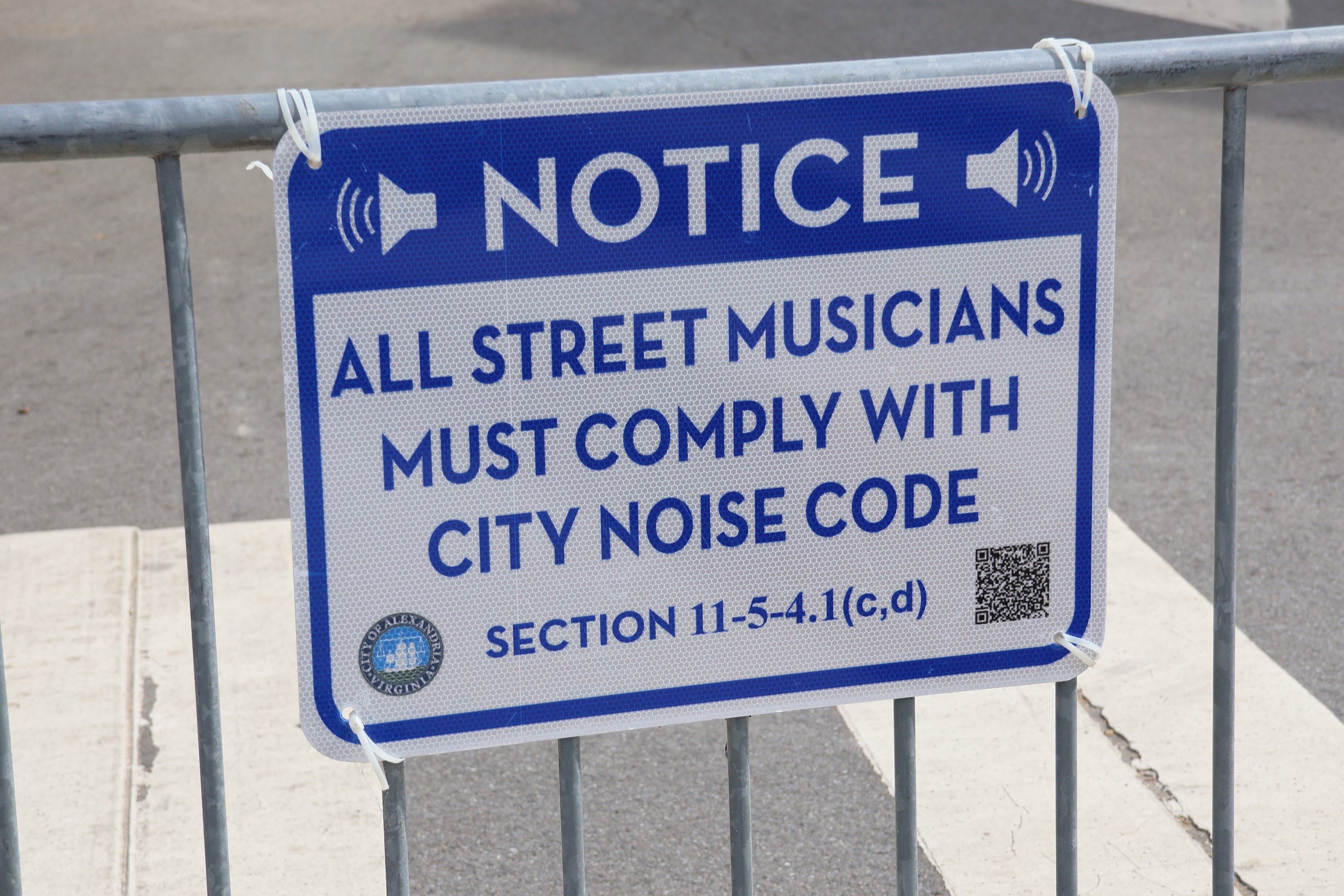 Anschlagtafel mit einem Schild, auf dem "Hinweis: Alle Straßenmusiker müssen die Stadtlärmvorschriften einhalten" steht, an einem Zaun befestigt, mit einer Straße im Hintergrund.